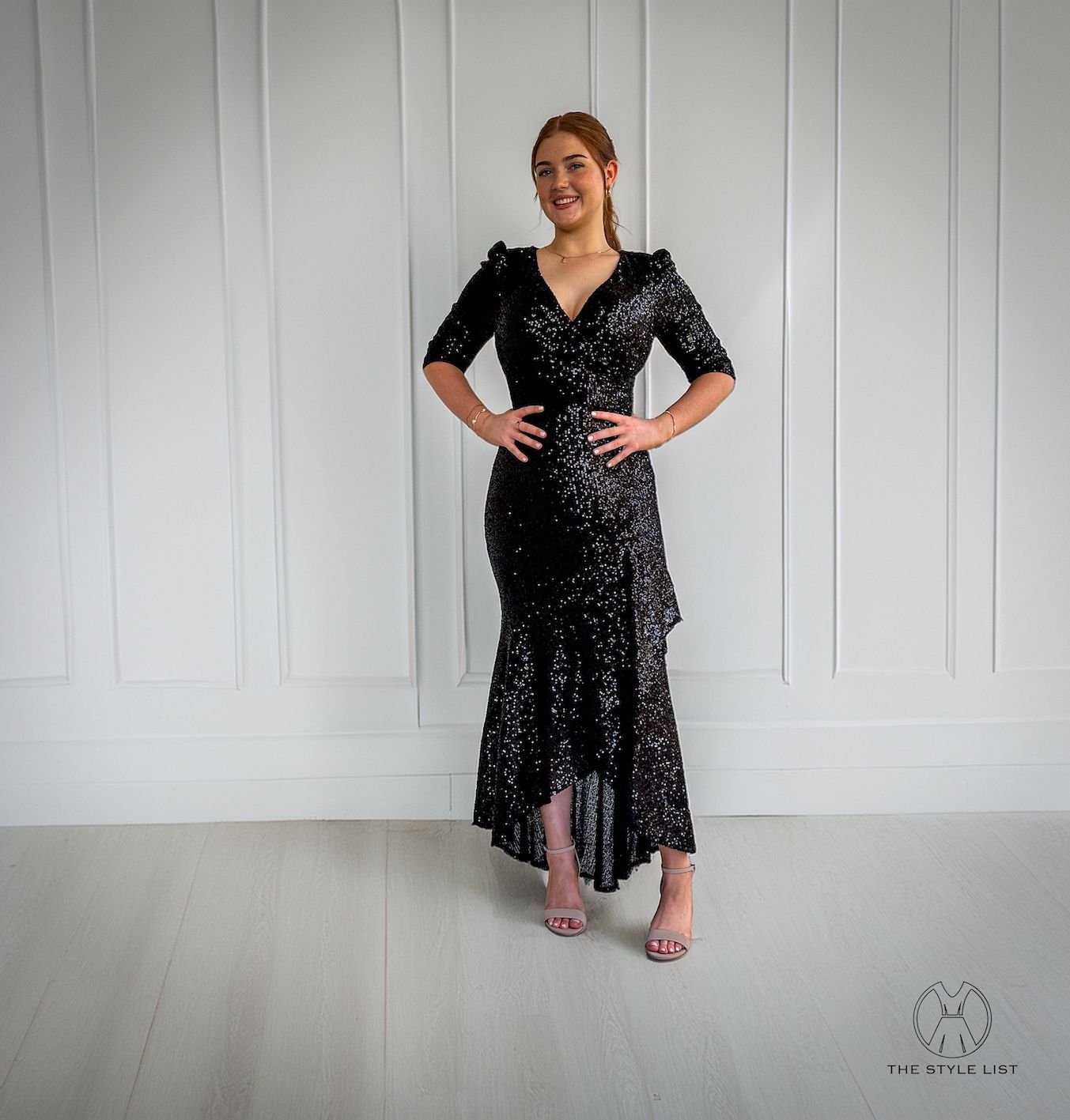 A woman in a black sequined dress is standing in front of a white wall.