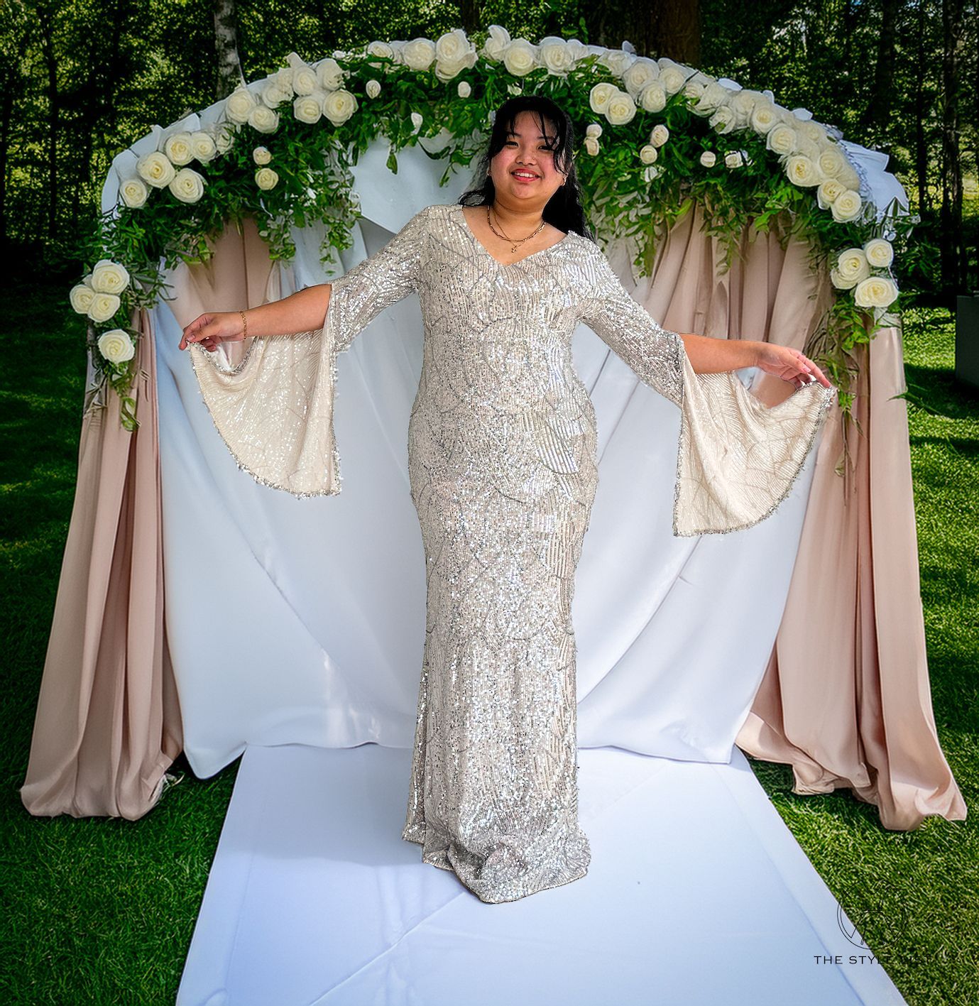 A woman in a sequined dress stands in front of a floral arch