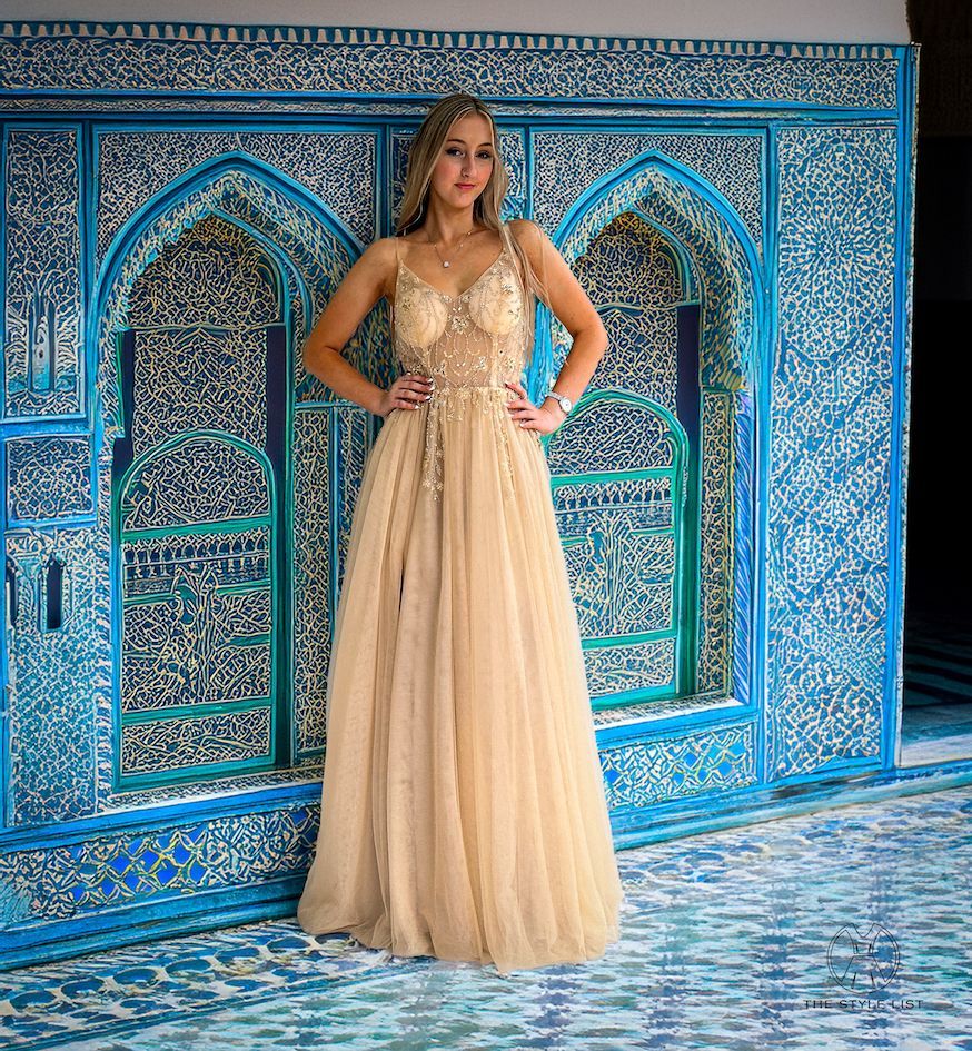 A woman in a long gold dress is standing in front of a blue wall.
