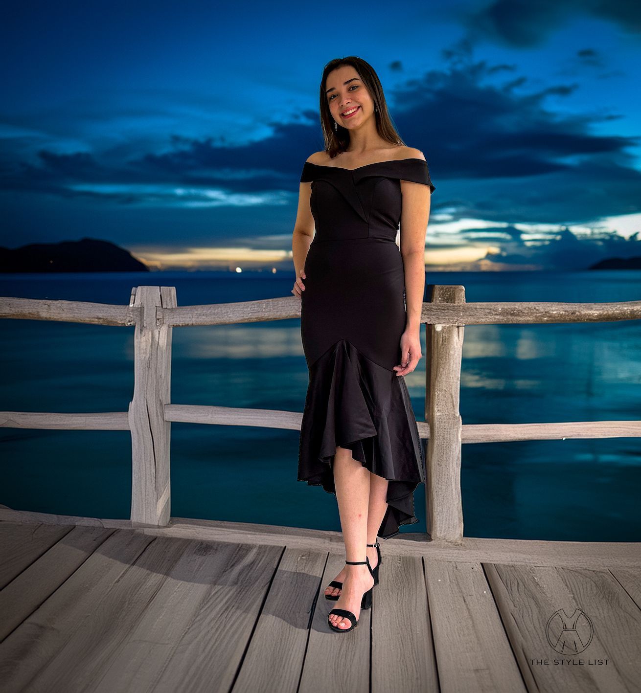 A woman in a black dress is standing on a pier overlooking the ocean