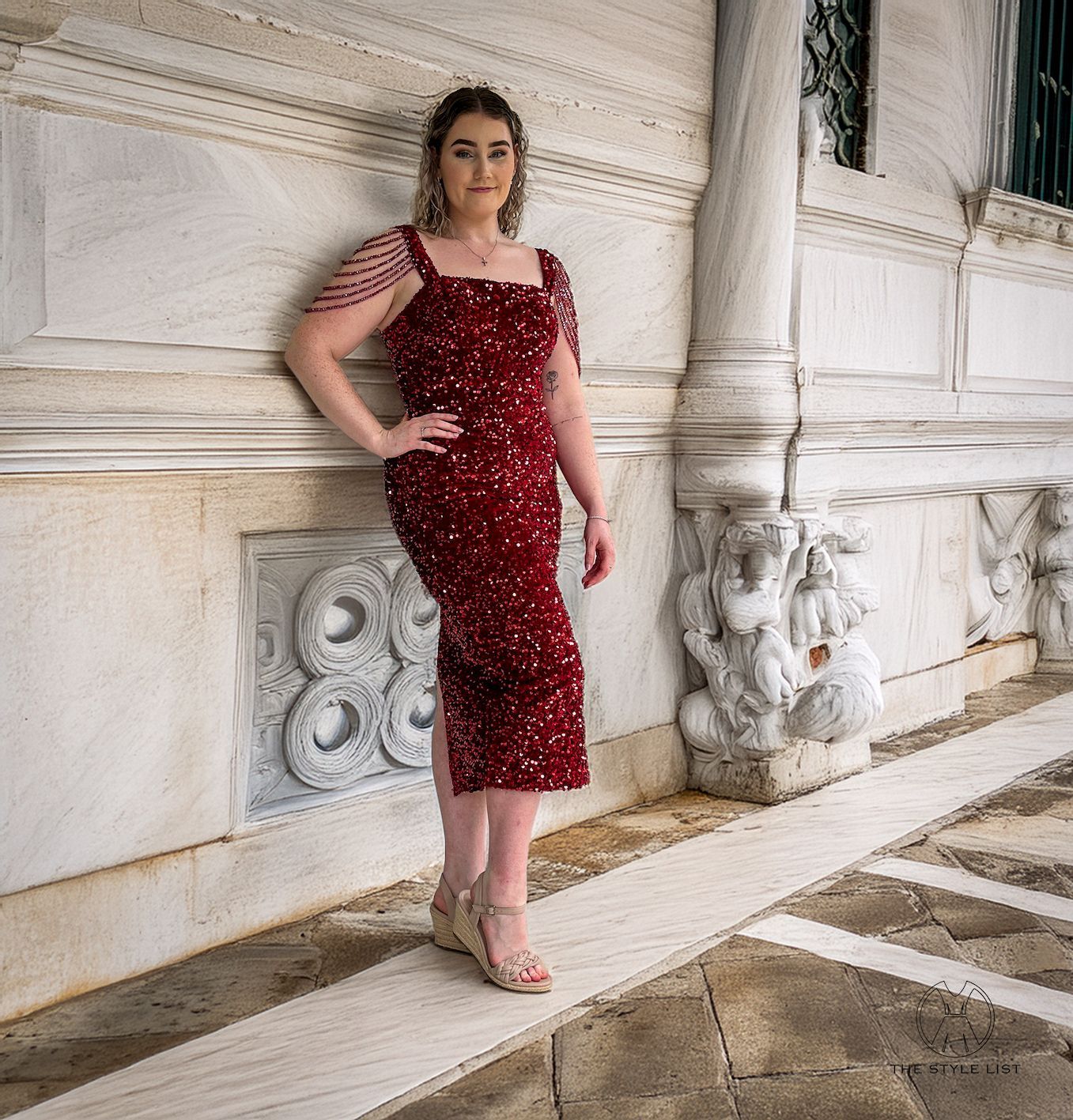 A woman in a red dress is leaning against a wall.