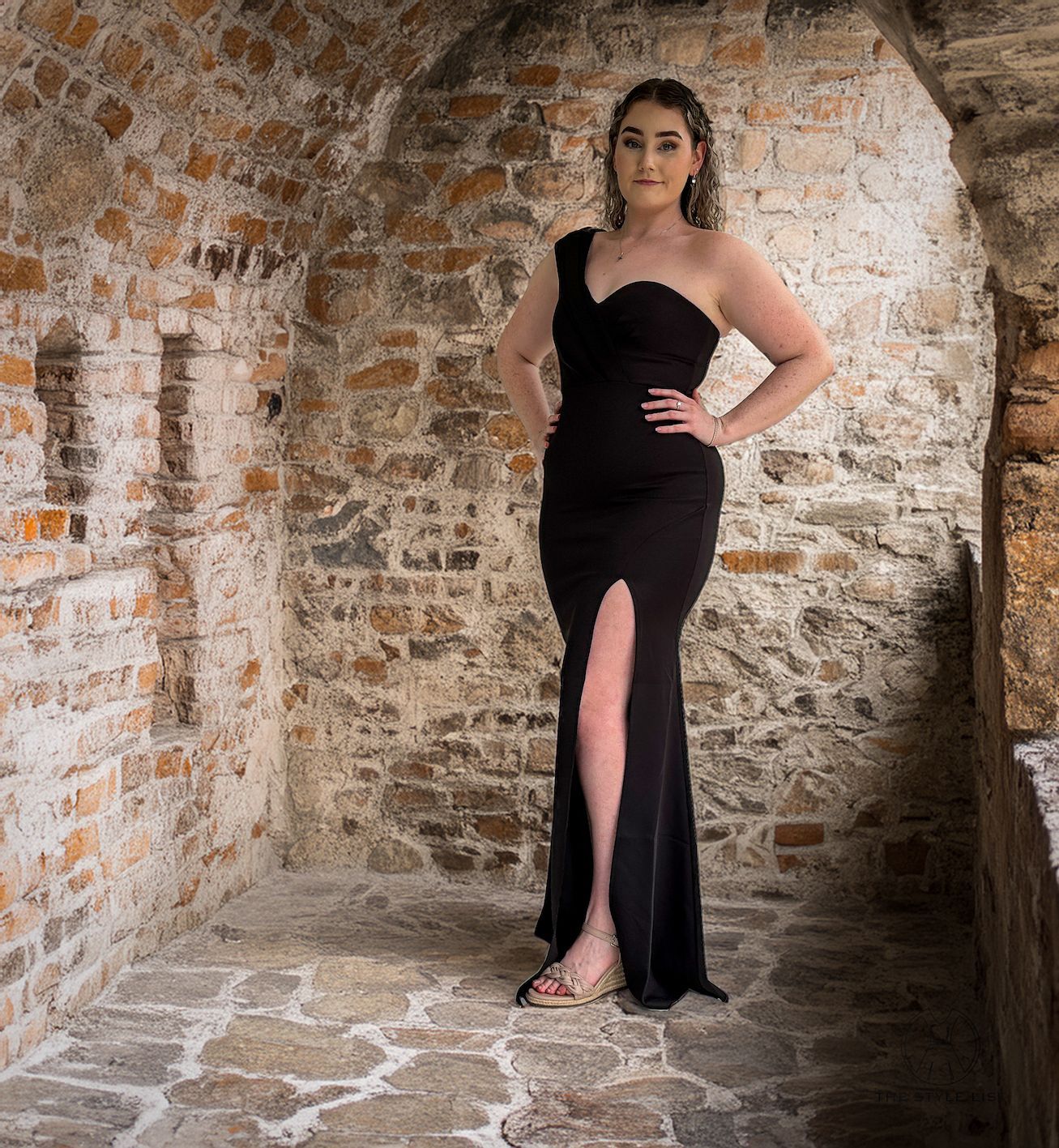 A woman in a black dress is standing in front of a brick wall.
