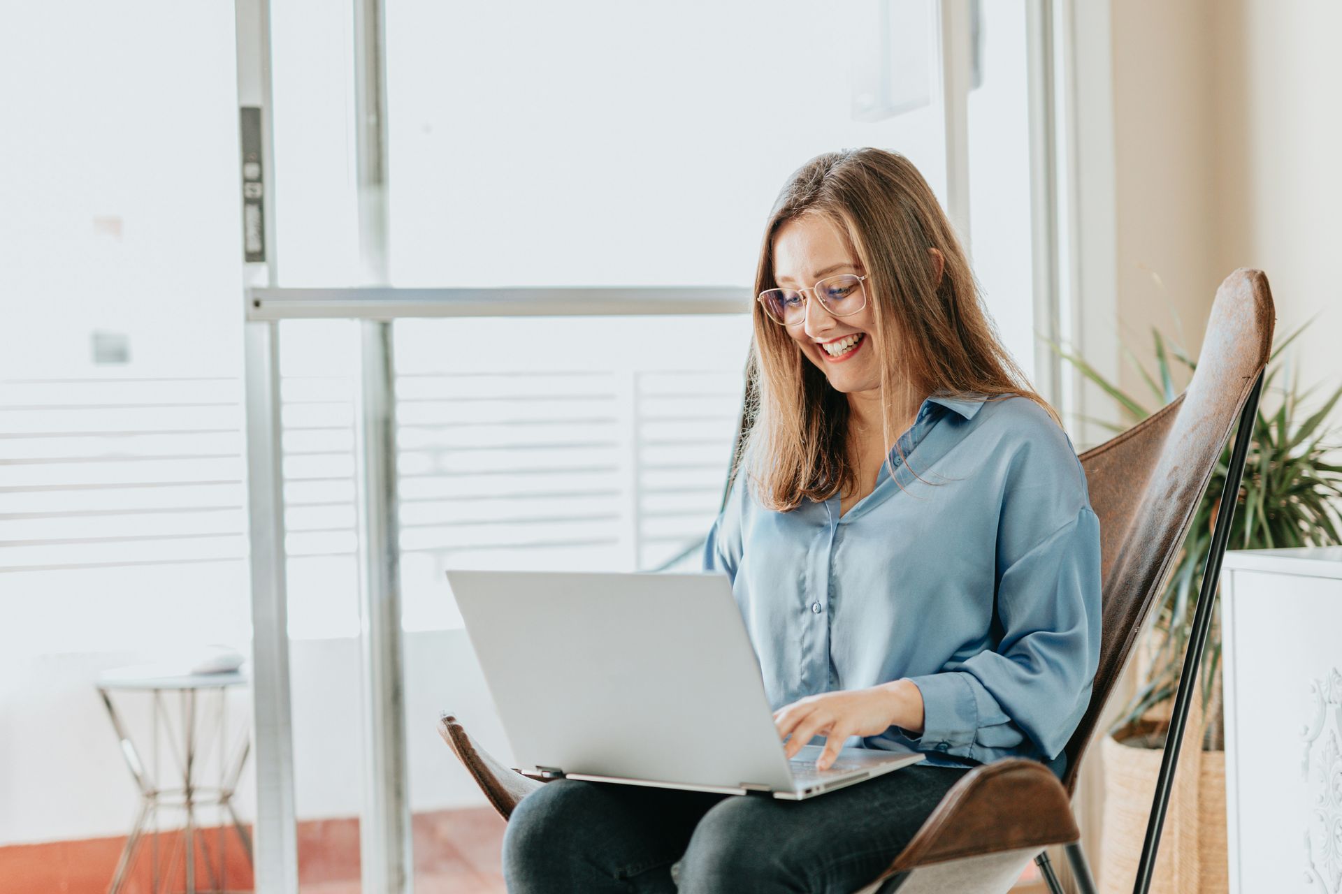 Woman in blue shirt smiles while using a laptop, seated in a chair near a window.