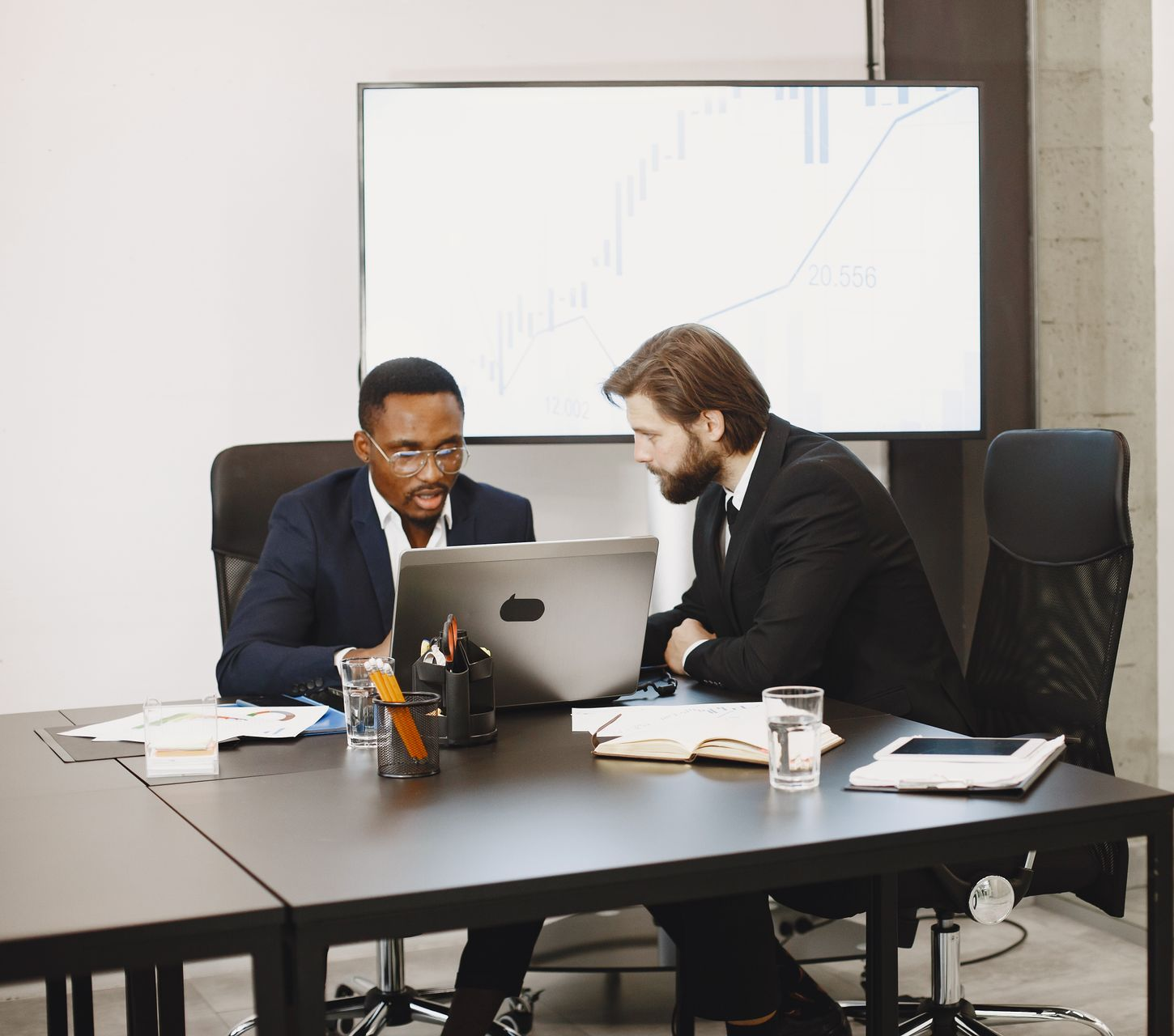 Two men in suits at a table, looking at a laptop with a graph on a screen in the background.