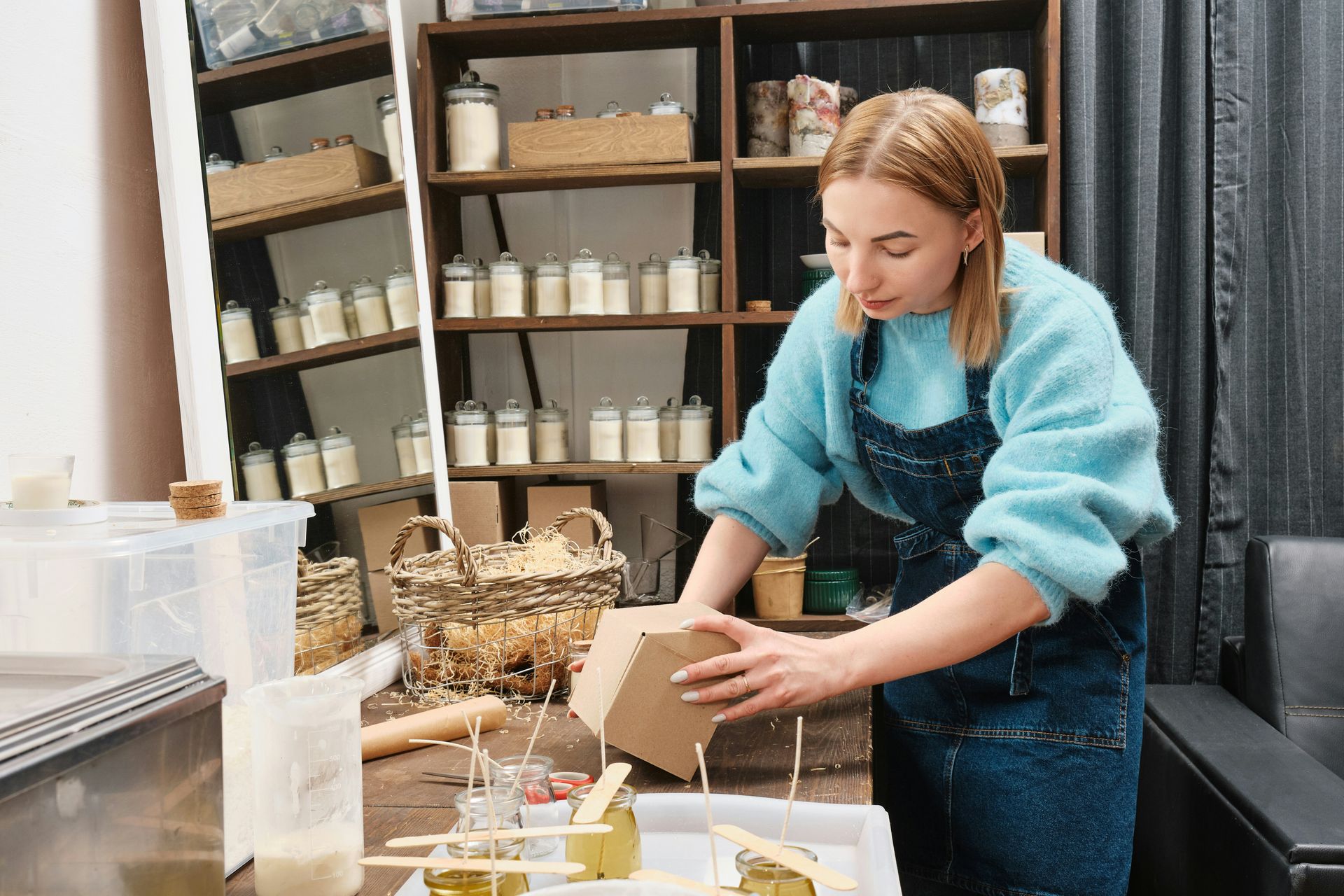 Woman in blue sweater and apron works with candles in a workshop.