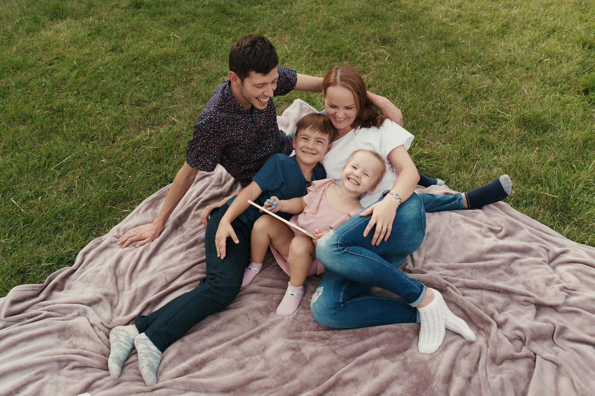 Family of four on a blanket in grass, smiling and looking at tablet.Icon
