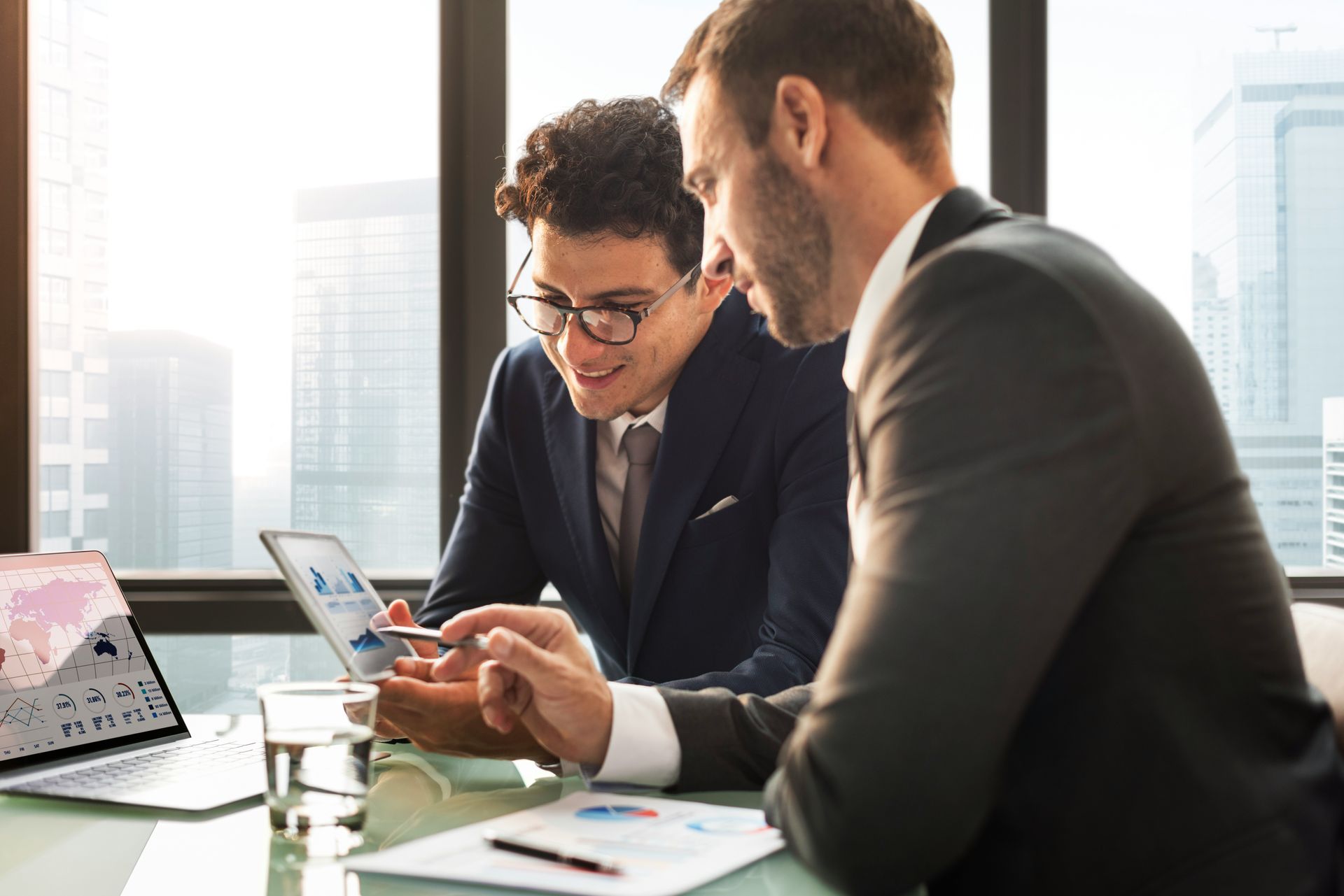 Two men in suits, collaborating over a tablet and papers, in an office with city views.Icon