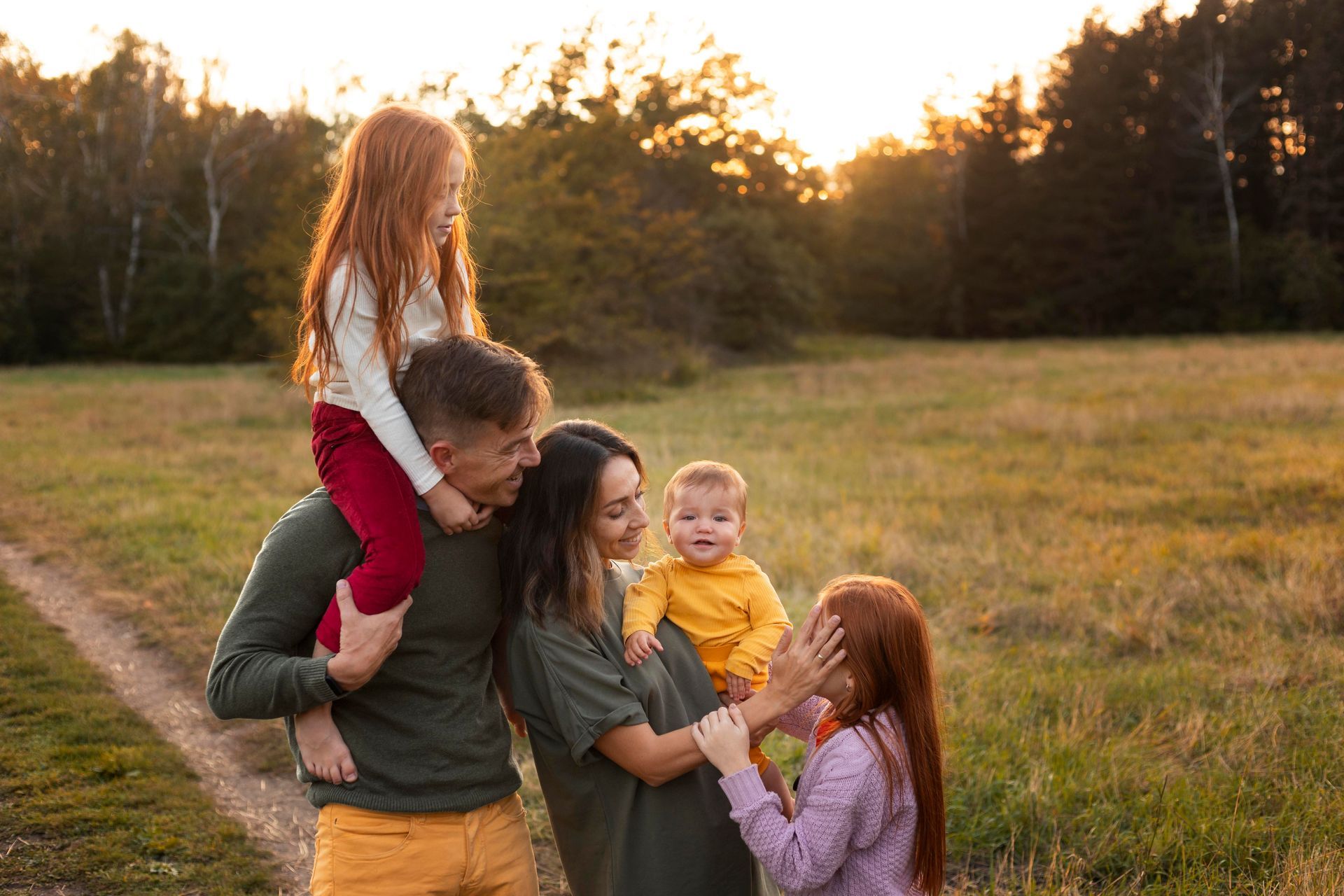 Family in a field at sunset; child on shoulders, others smile and play, warm light.