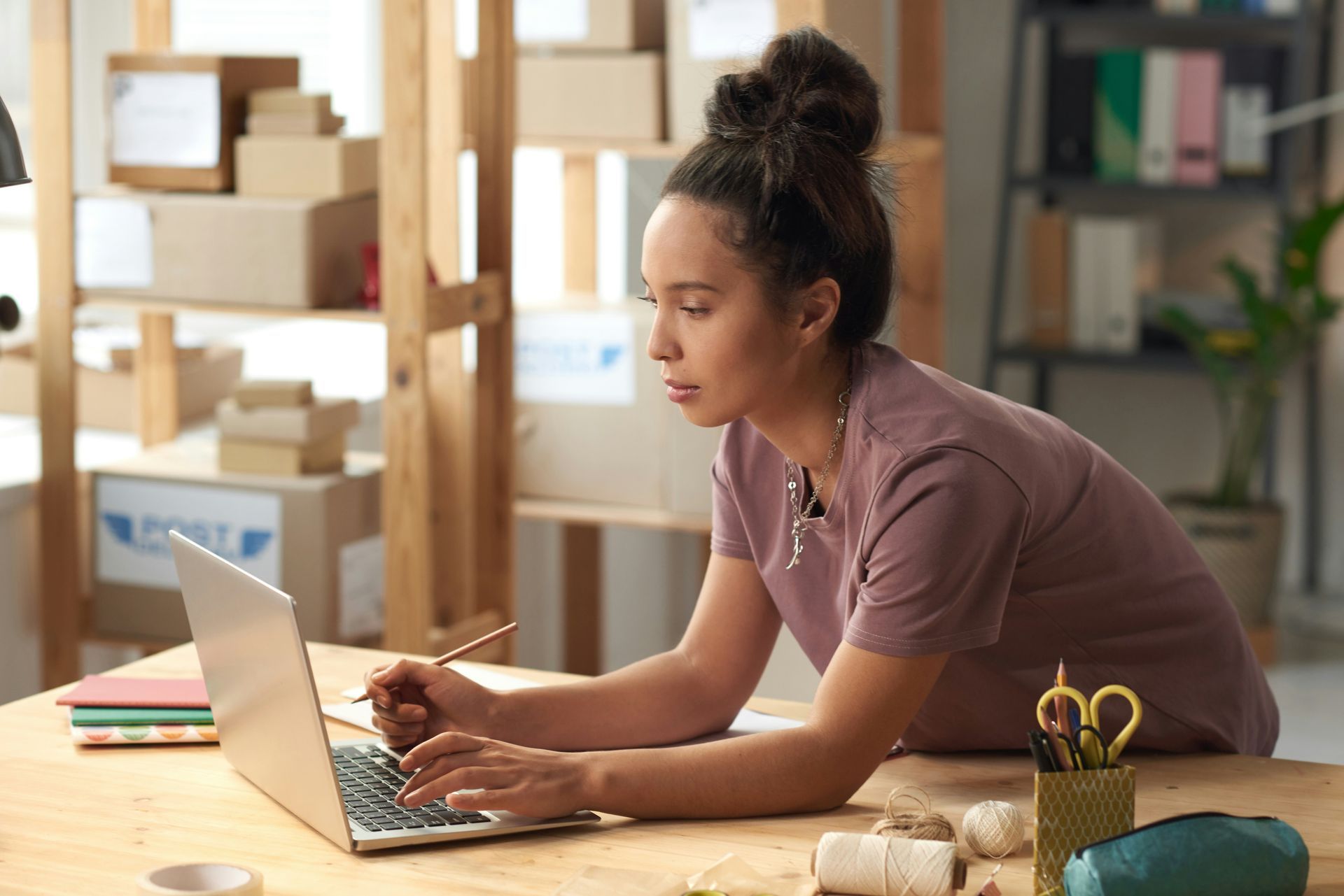 Woman working on laptop at a wooden desk with craft supplies, surrounded by boxes on shelves.