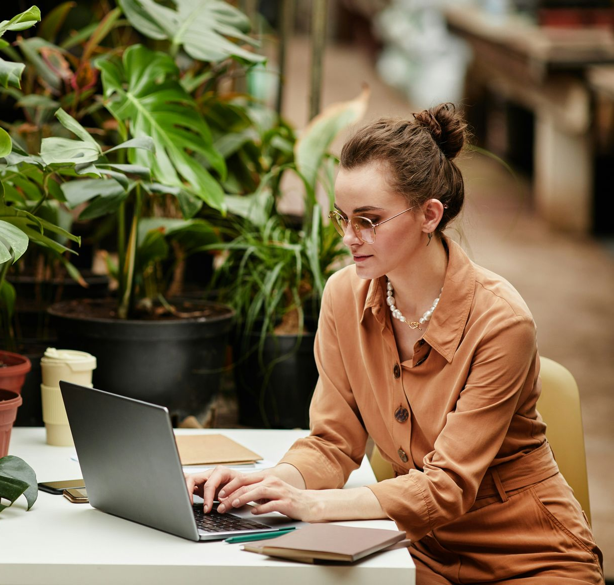 Woman in brown outfit typing on laptop at a table surrounded by plants.Icon