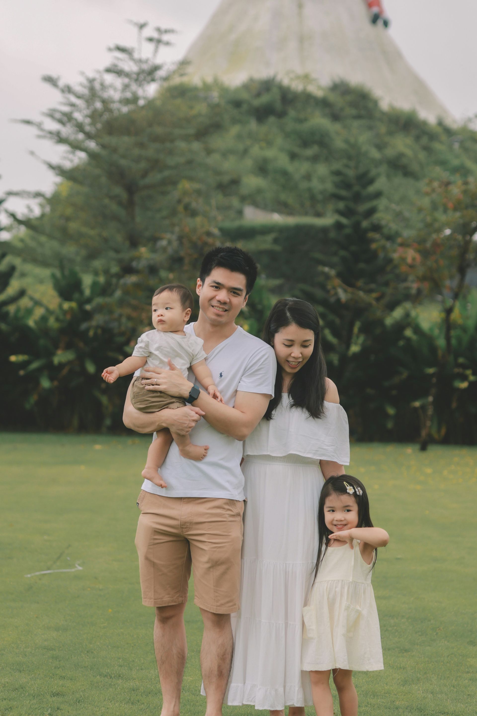 Family of four smiling on grass in front of a unique structure and trees.