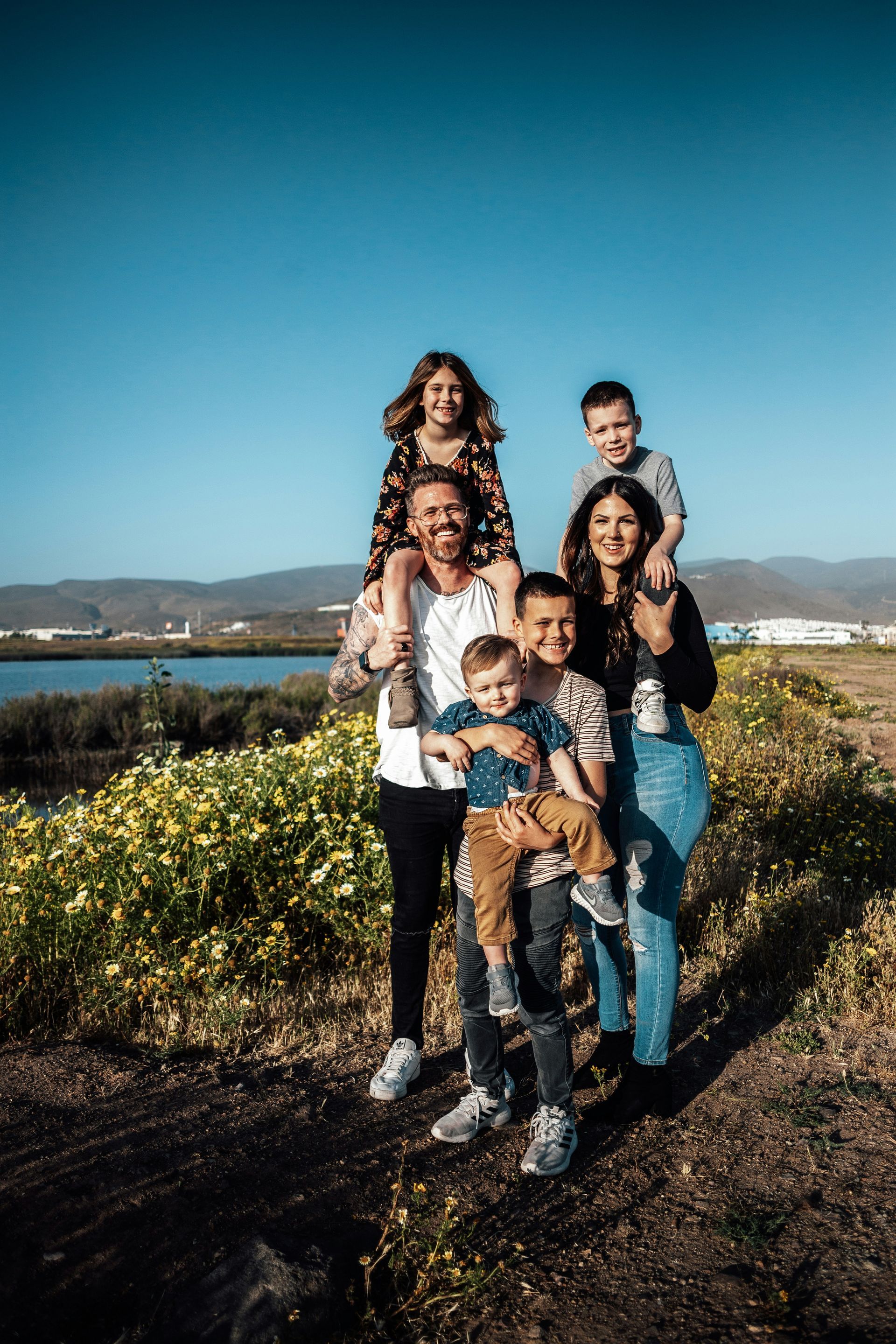 Family of six posing outdoors with two children on shoulders. Sunny day, yellow flowers.