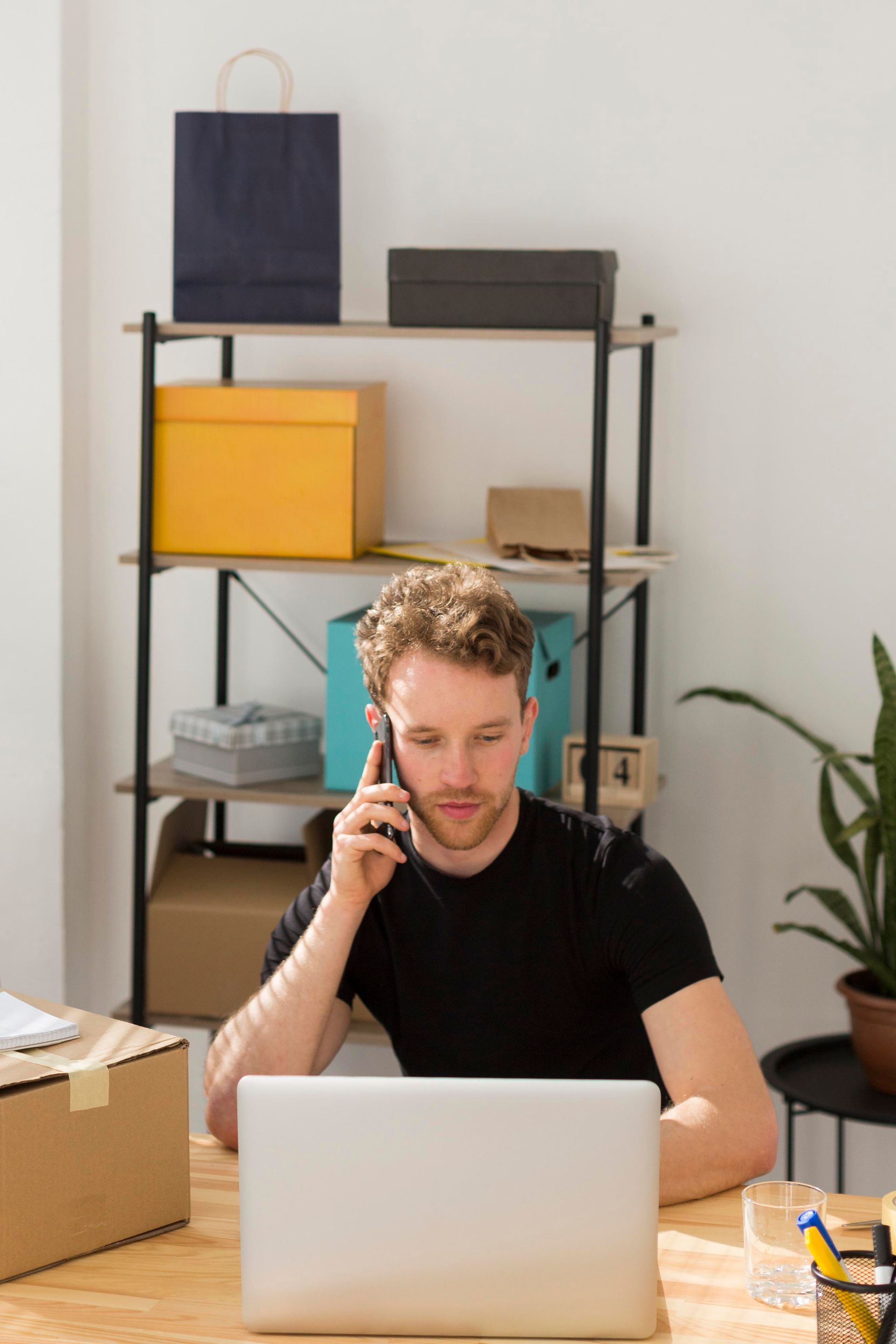 Man on phone, looking at laptop, seated at desk with shelving holding boxes and supplies.
