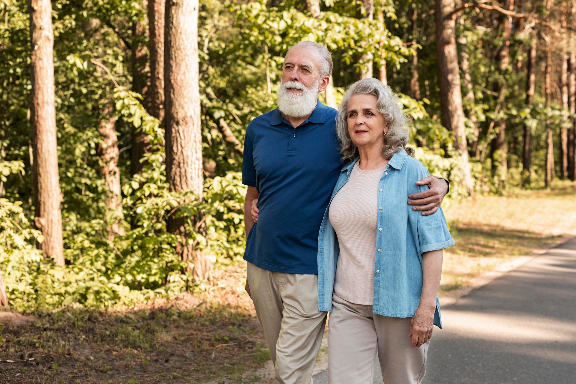 Couple in forest, man with white beard and blue shirt, woman with curly hair and denim shirt, arm in arm.