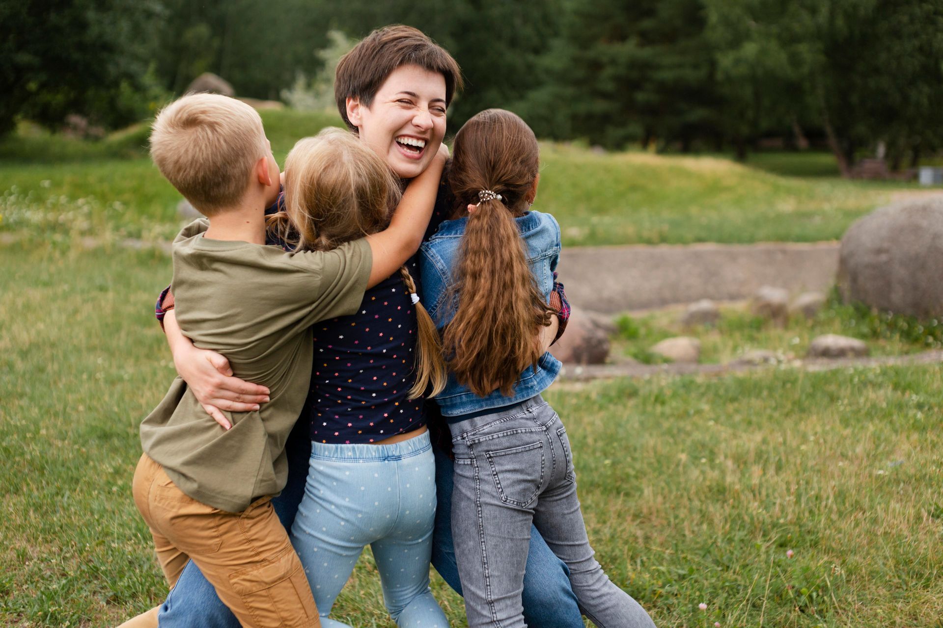 Woman laughing, embraced by three children in a grassy park setting.