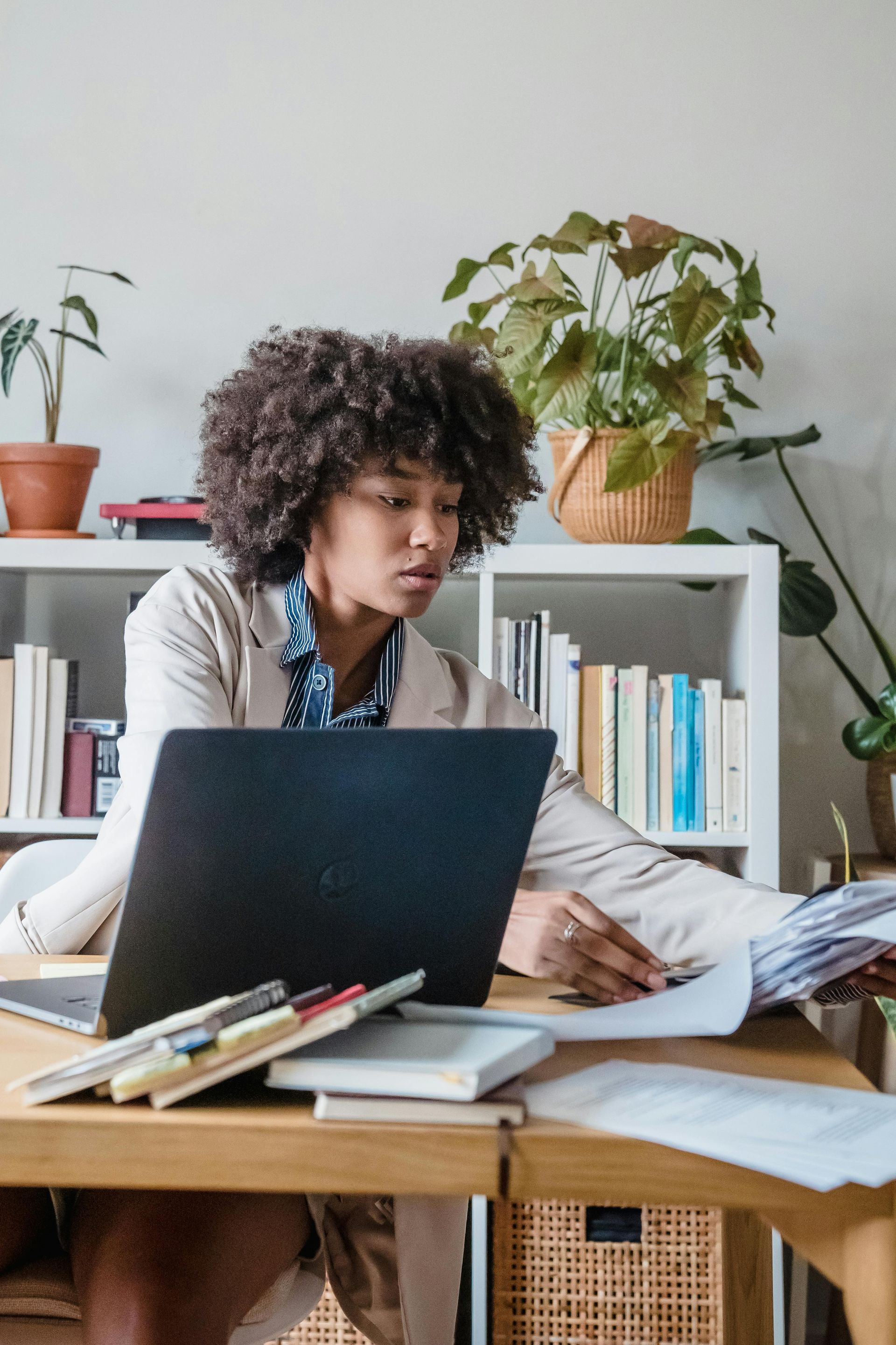 Woman working at desk with laptop, reviewing documents, surrounded by plants and bookshelves.Icon