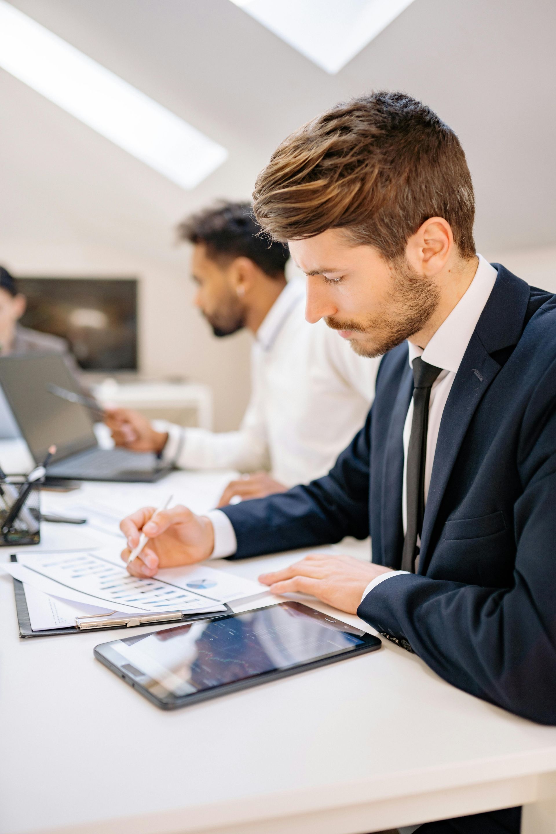 Man in suit reviewing charts with a pen, another man in background working on a laptop.