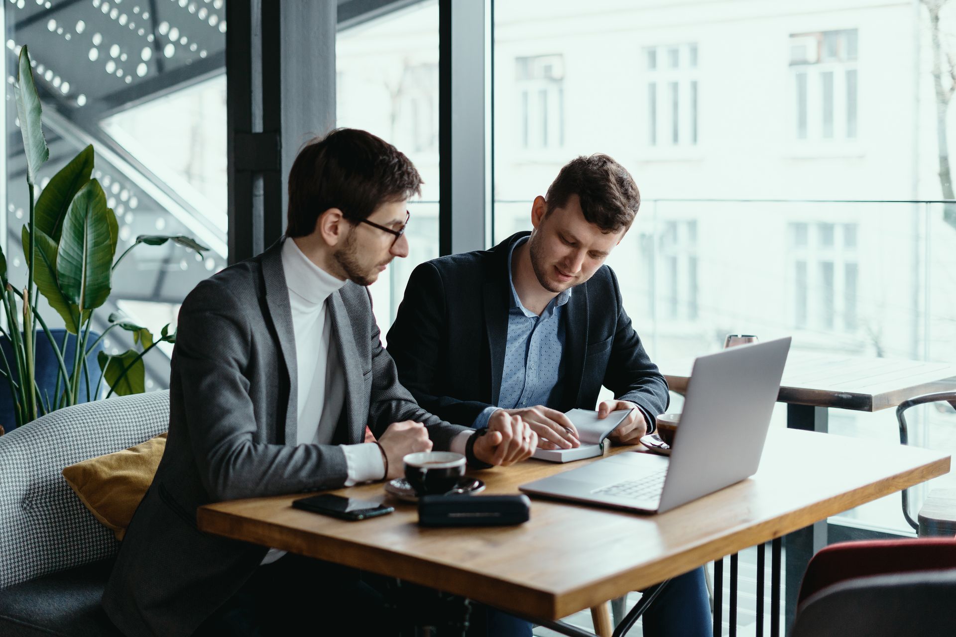 Two men in suits at a table with a laptop and papers, likely discussing business in a cafe.