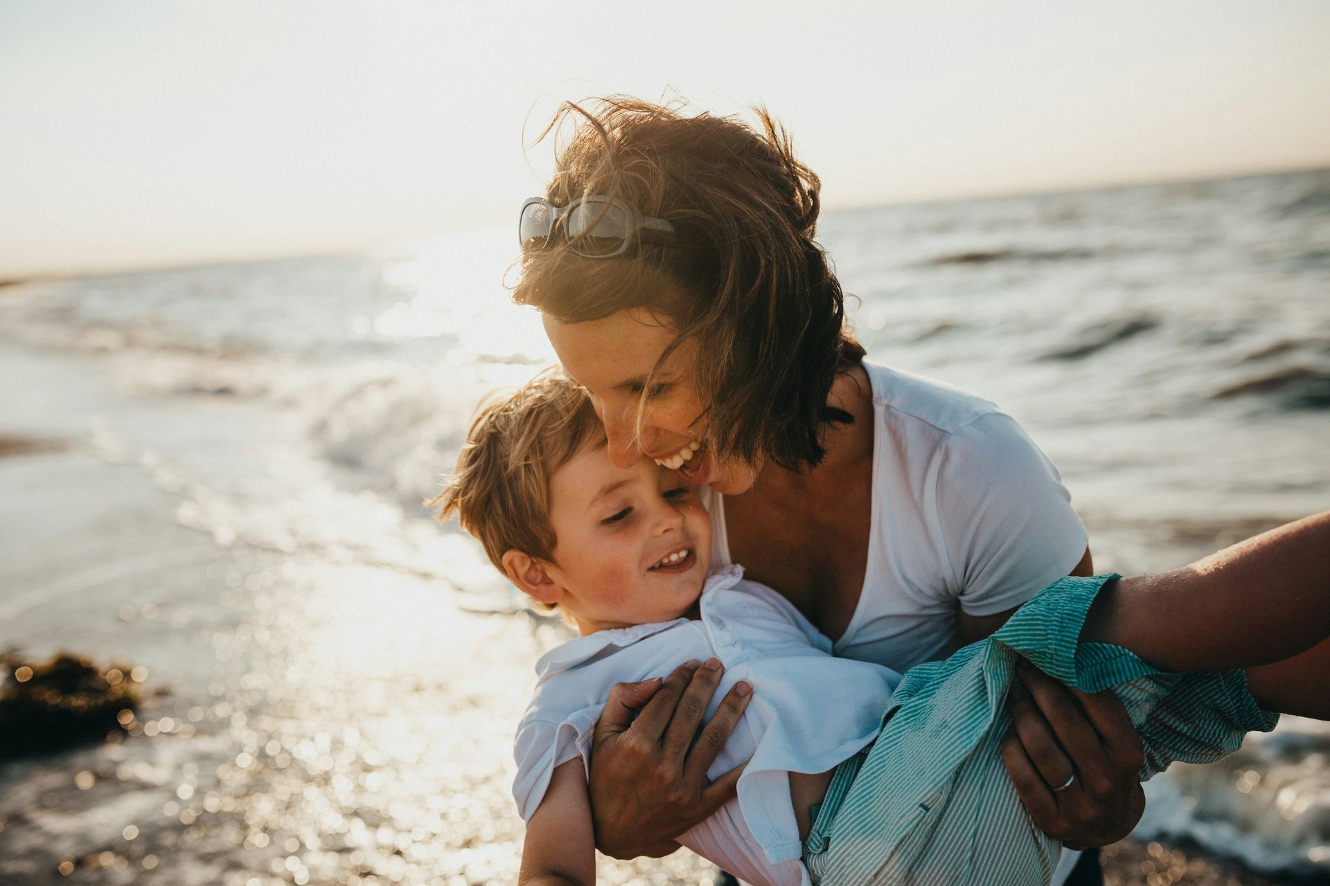 Woman holding and kissing a child on the beach; golden sunlight, ocean waves, happy expressions.