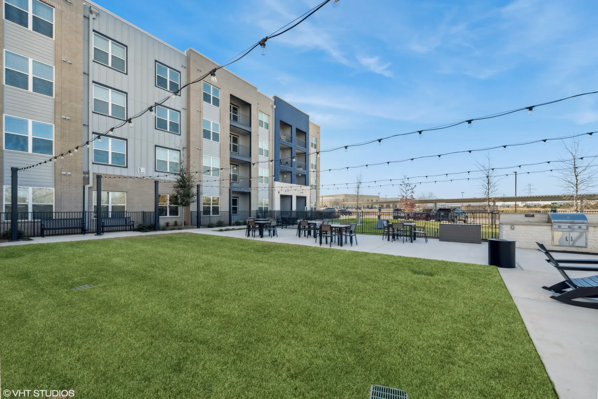 Outdoor community courtyard with string lights, tables, and grass next to an apartment building.