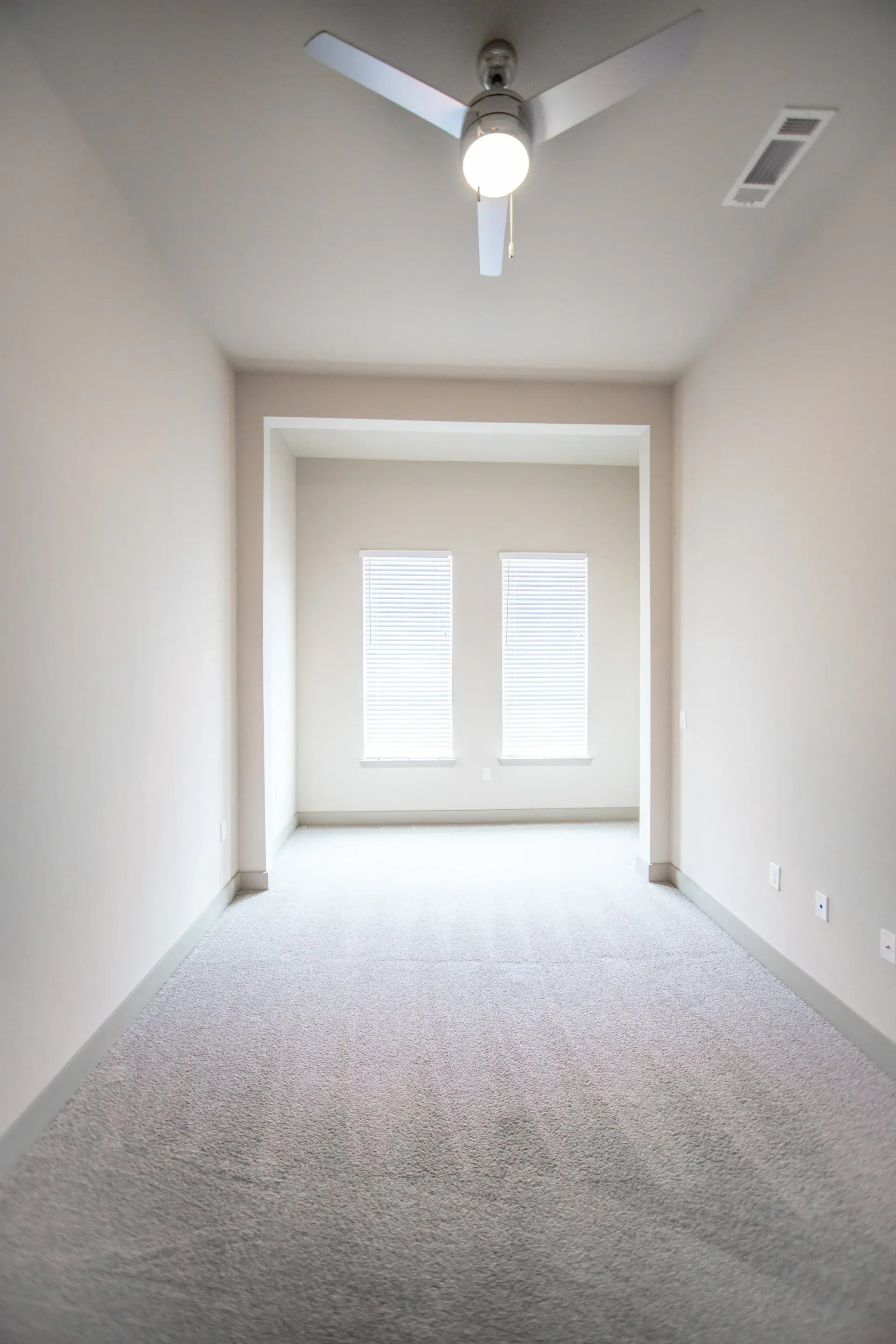 Empty carpeted room with a ceiling fan and two tall windows with blinds at the far end.