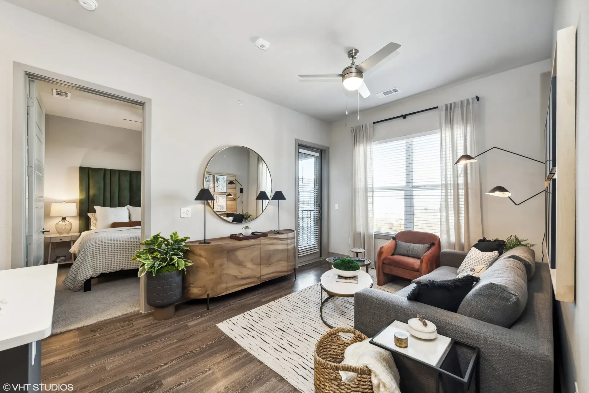 Living room of a modern apartment with a gray sofa, round mirror, wood-toned console, and a large window with sheer curtains.