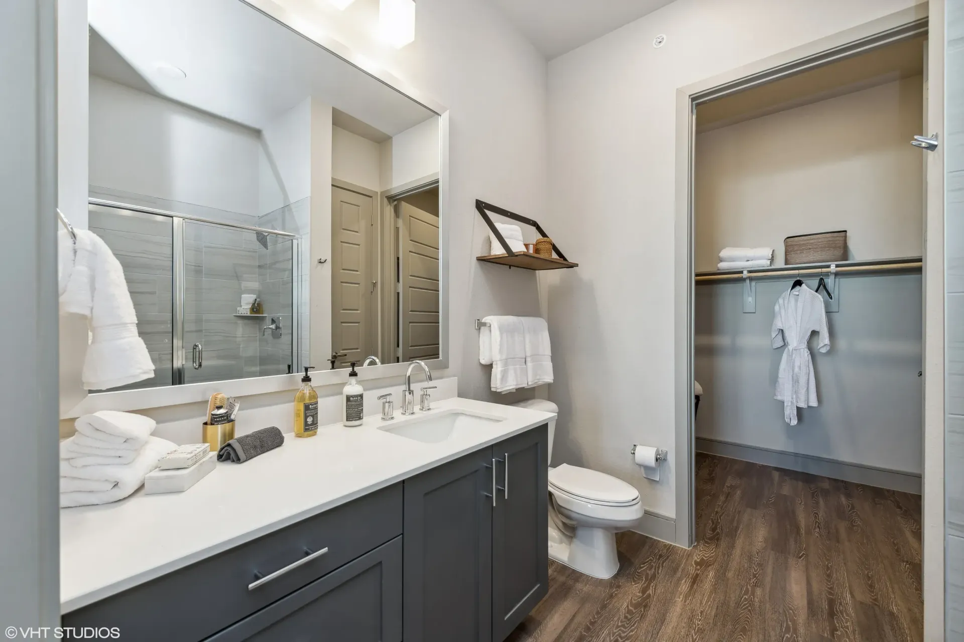 Modern apartment bathroom with double vanity, large mirror, glass shower, toilet, and open closet.