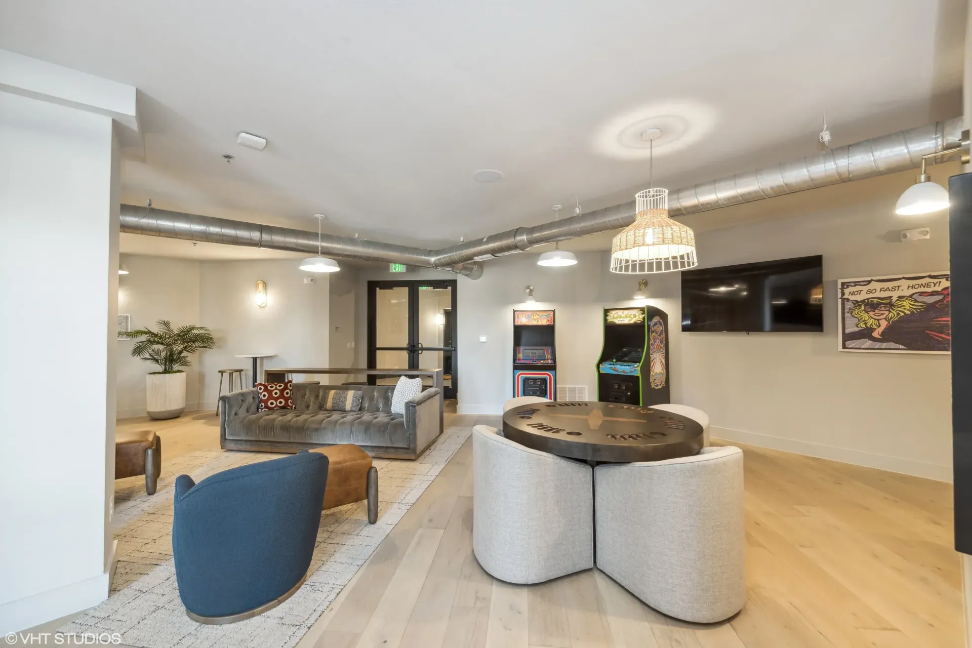 Indoor community lounge with sofas, arcade games, a round table, and a wall-mounted TV.