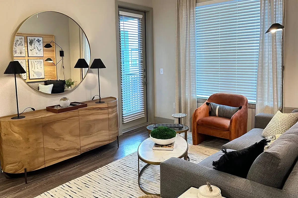 Living room interior with a round wall mirror, wooden console, grey sofa, orange chair, and window blinds.