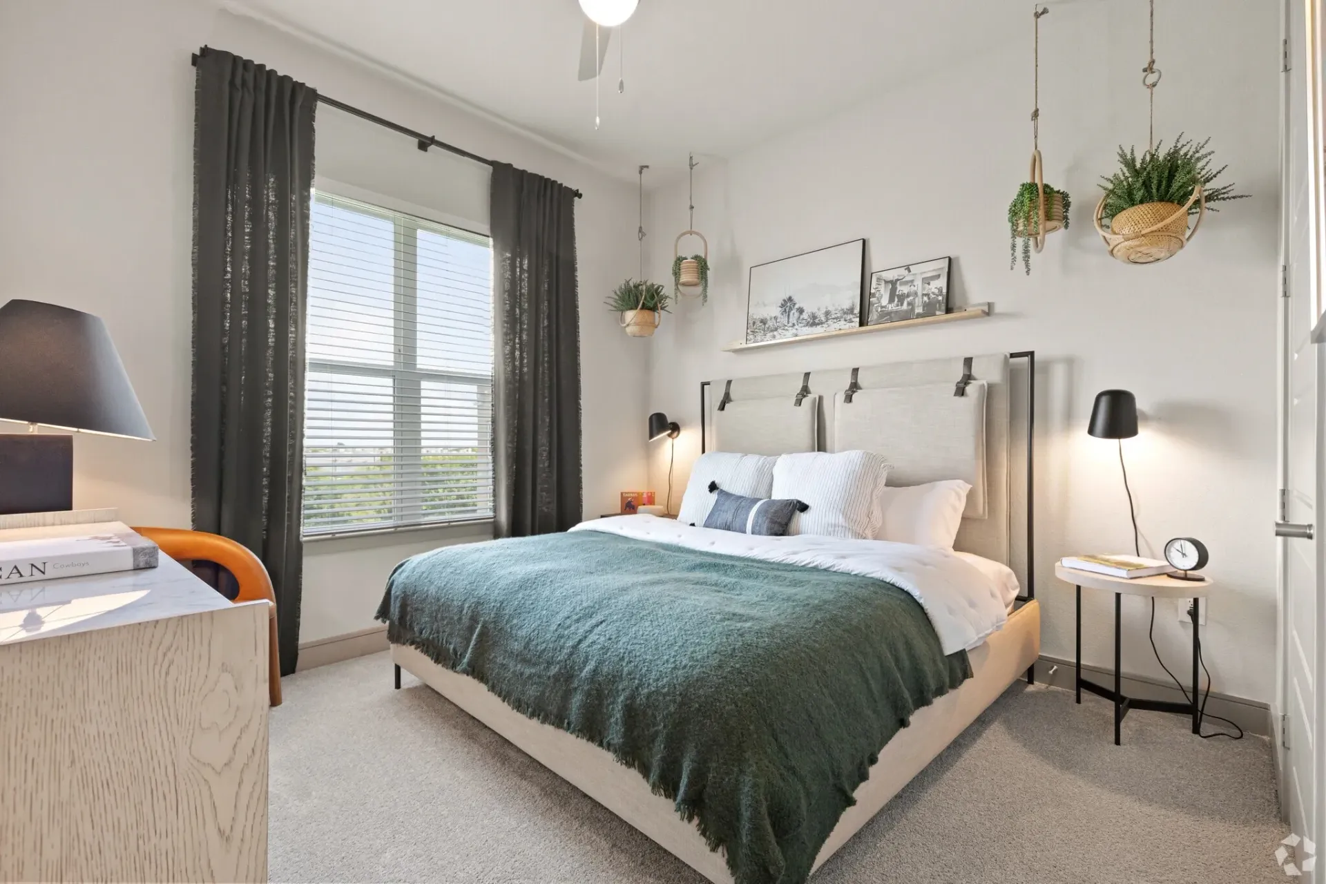 Bright apartment bedroom with a large window, bed, nightstands, and hanging plants.