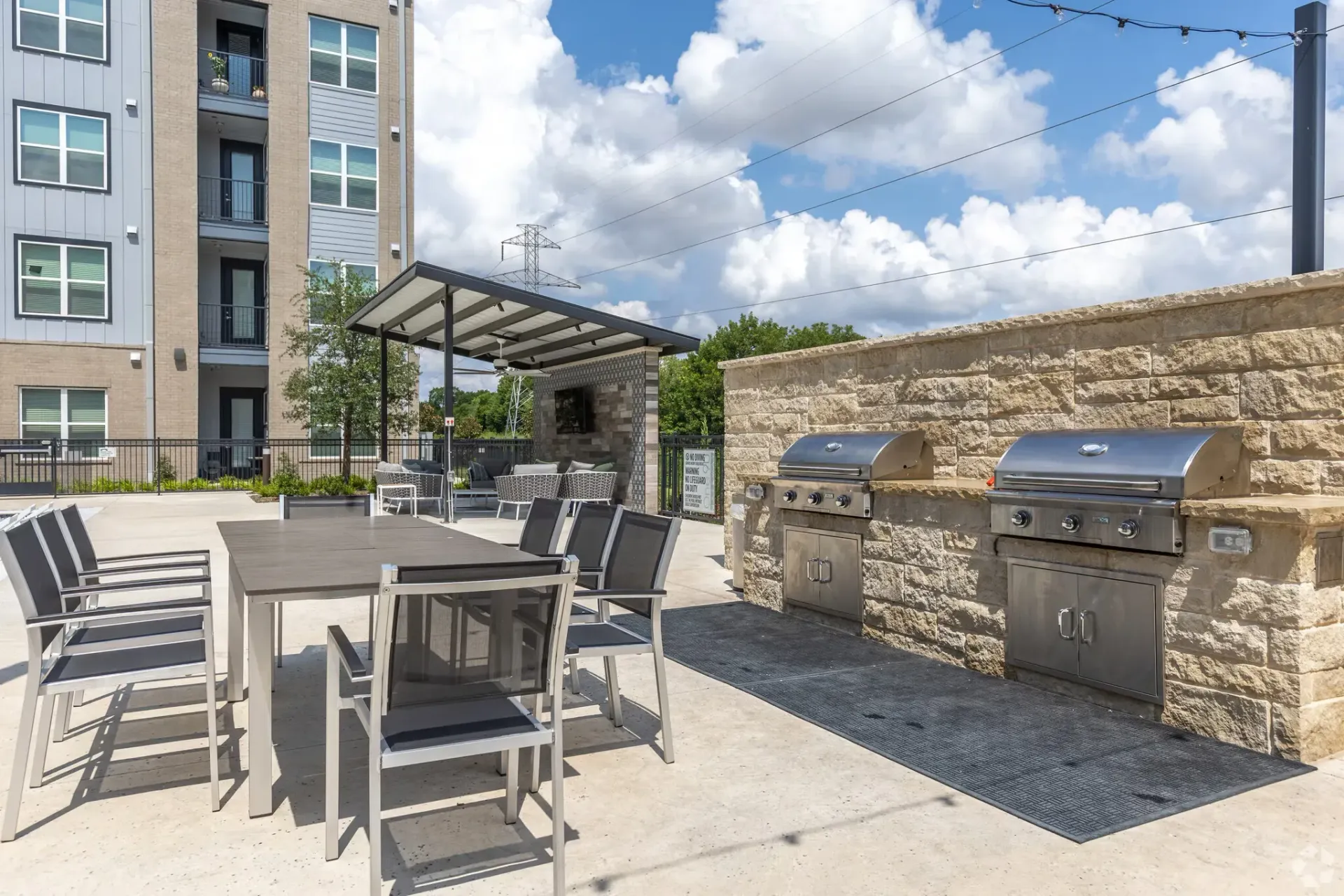 Outdoor communal amenity with grills, tables, and a covered seating pavilion beside a modern apartment building.