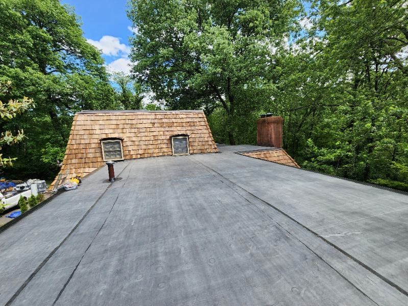 Roof partially covered with black material and cedar shingles. Chimney and windows are visible, set in a leafy environment.