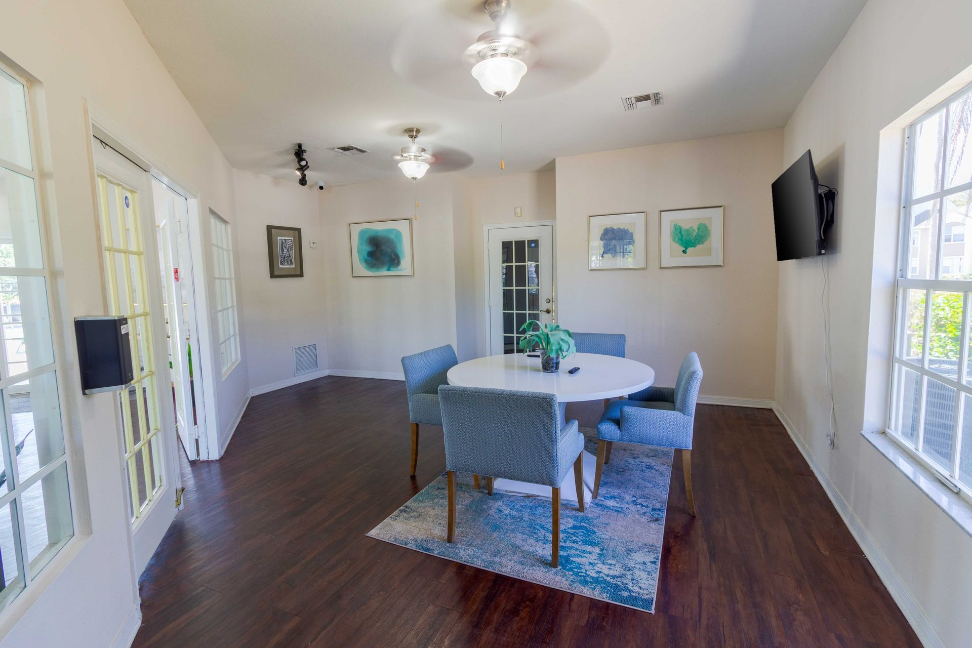 Dining room with round table, four blue chairs, artwork, TV, and windows at The Point at Naples in Naples, FL.