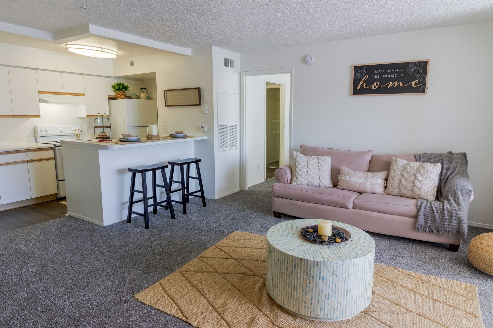 Open-plan living space: kitchen with stools, blush sofa, round coffee table on a rug; gray carpet at The Point at Naples in Naples, FL.