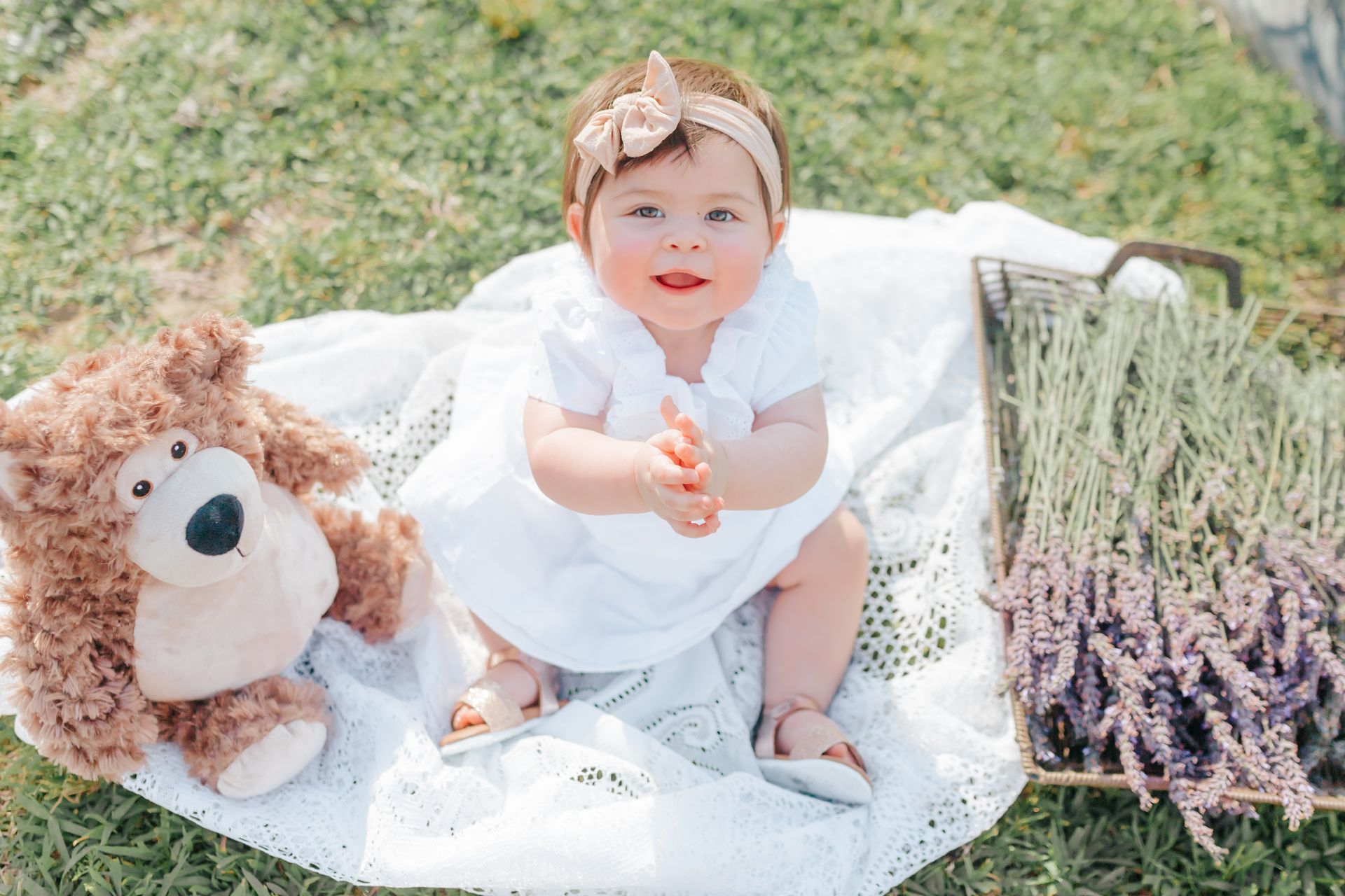 A baby girl is sitting on a blanket next to a teddy bear.