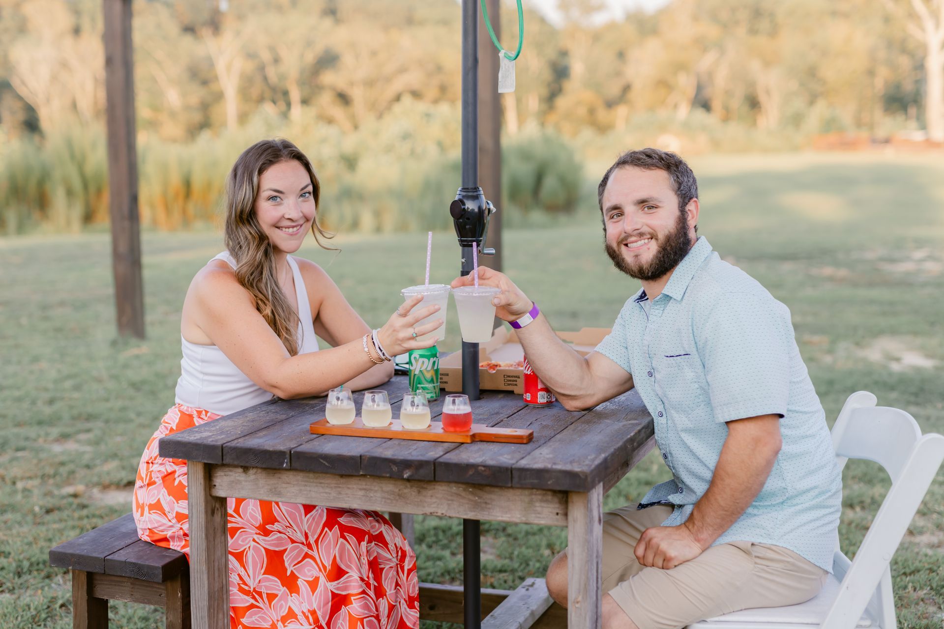 A man and a woman are sitting at a picnic table toasting with drinks.