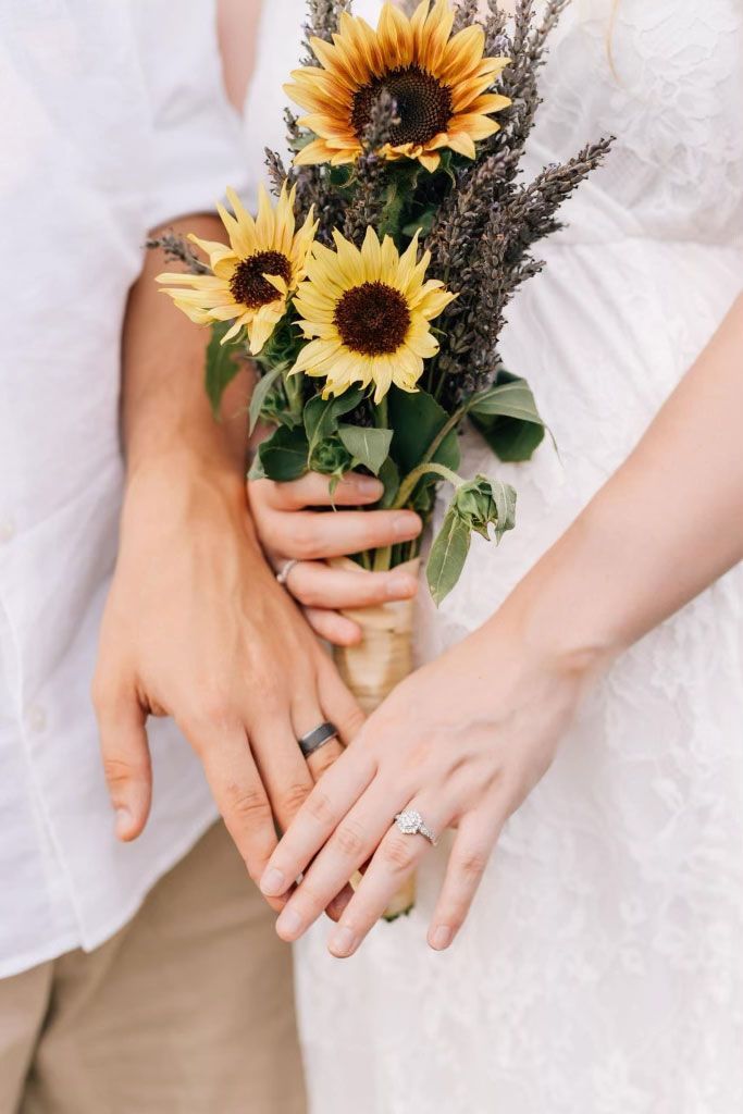 A bride and groom are holding hands while holding a bouquet of sunflowers.