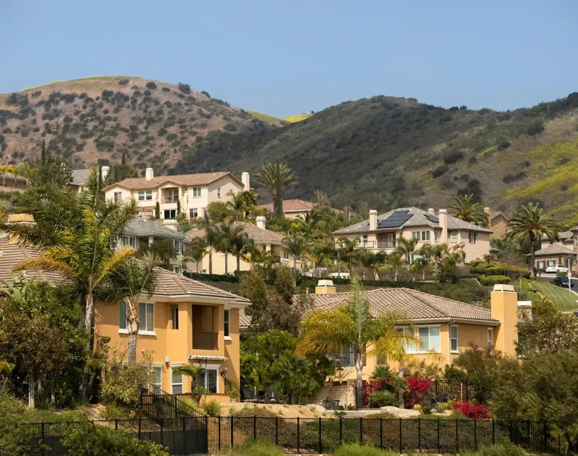 Stucco houses nestled on a hillside with dry, grassy slopes and scattered trees under a clear blue sky.