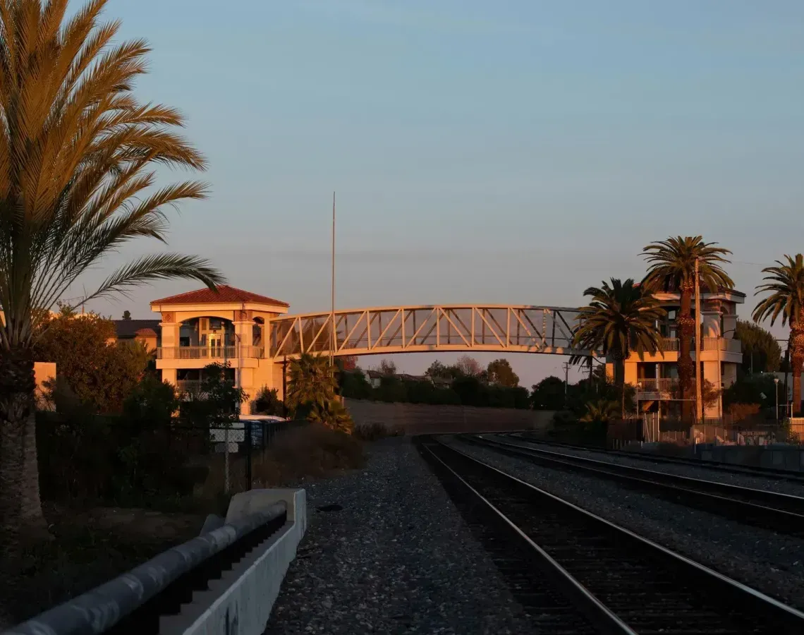 A pedestrian bridge connects two buildings over a set of railroad tracks during a golden sunset.