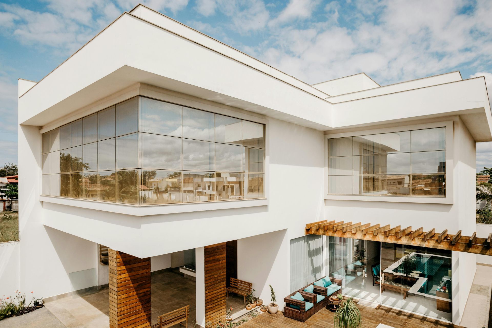 A modern two-story white house with large glass windows, wood-paneled pillars, and a wooden pergola over a patio area.