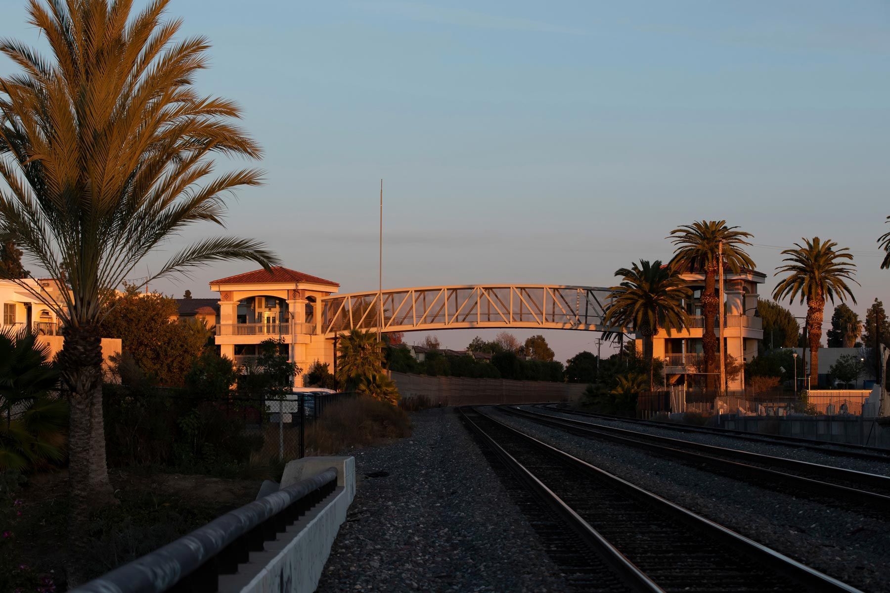 A pedestrian bridge connects two buildings over a set of railroad tracks during a golden sunset.