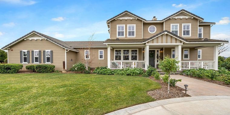A large, tan-colored two-story suburban home with a front porch, manicured lawn, and a clear blue sky background.