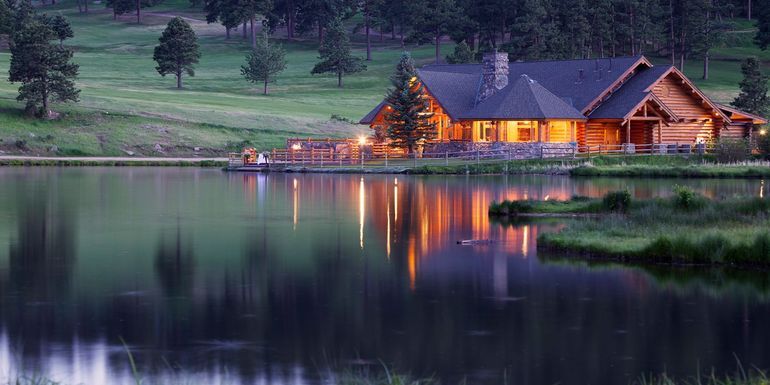 A rustic log cabin illuminated at dusk, nestled on the edge of a calm lake with trees in the background.