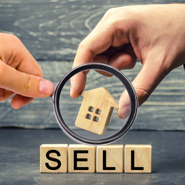 Two hands hold a magnifying glass over a small wooden house figure resting above wooden blocks that spell 