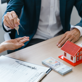 A person hands a house key to another across a desk with a contract, a stack of cash, and a small house model.