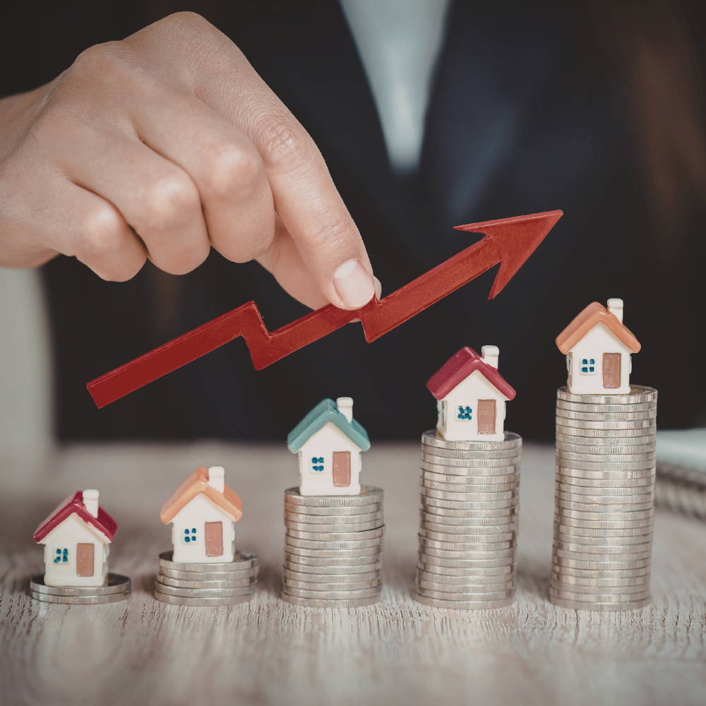 A hand holds a red upward-pointing arrow over five small house models atop increasing stacks of silver coins.
