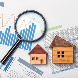 A magnifying glass rests over a bar chart, positioned beside two small wooden house models on a financial spreadsheet.