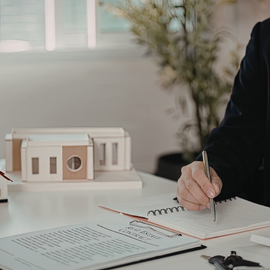 A professional works at a desk, sketching on a notepad next to a small architectural model in a brightly lit office.