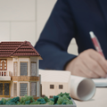 A model of a house sits on a desk in the foreground while a person writes on blueprints in the blurred background.