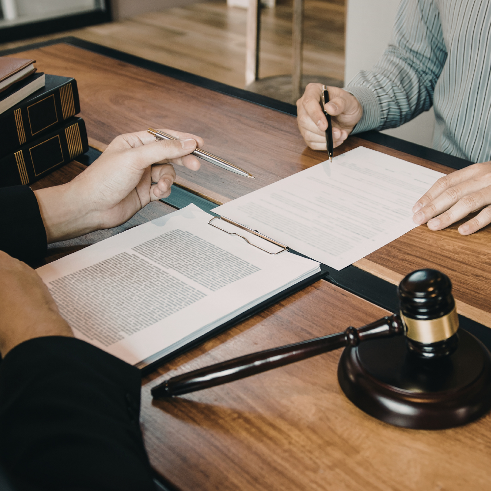 Two people review legal documents at a desk with a judge's gavel and law books, indicating a professional legal meeting.